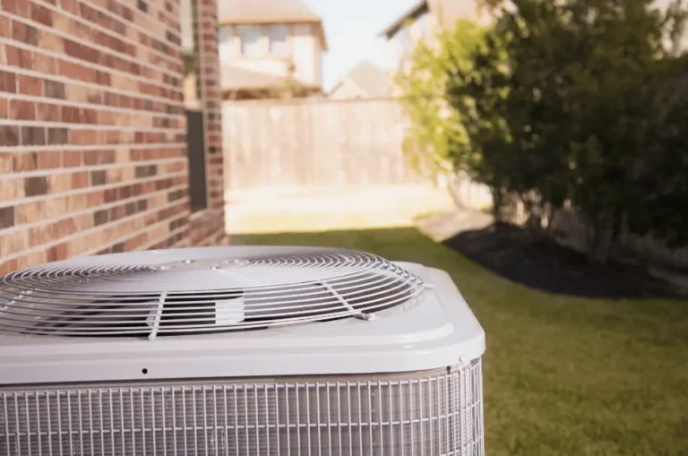 Outdoor air conditioner condenser unit beside a brick house, with a green lawn and shrubbery in the background.