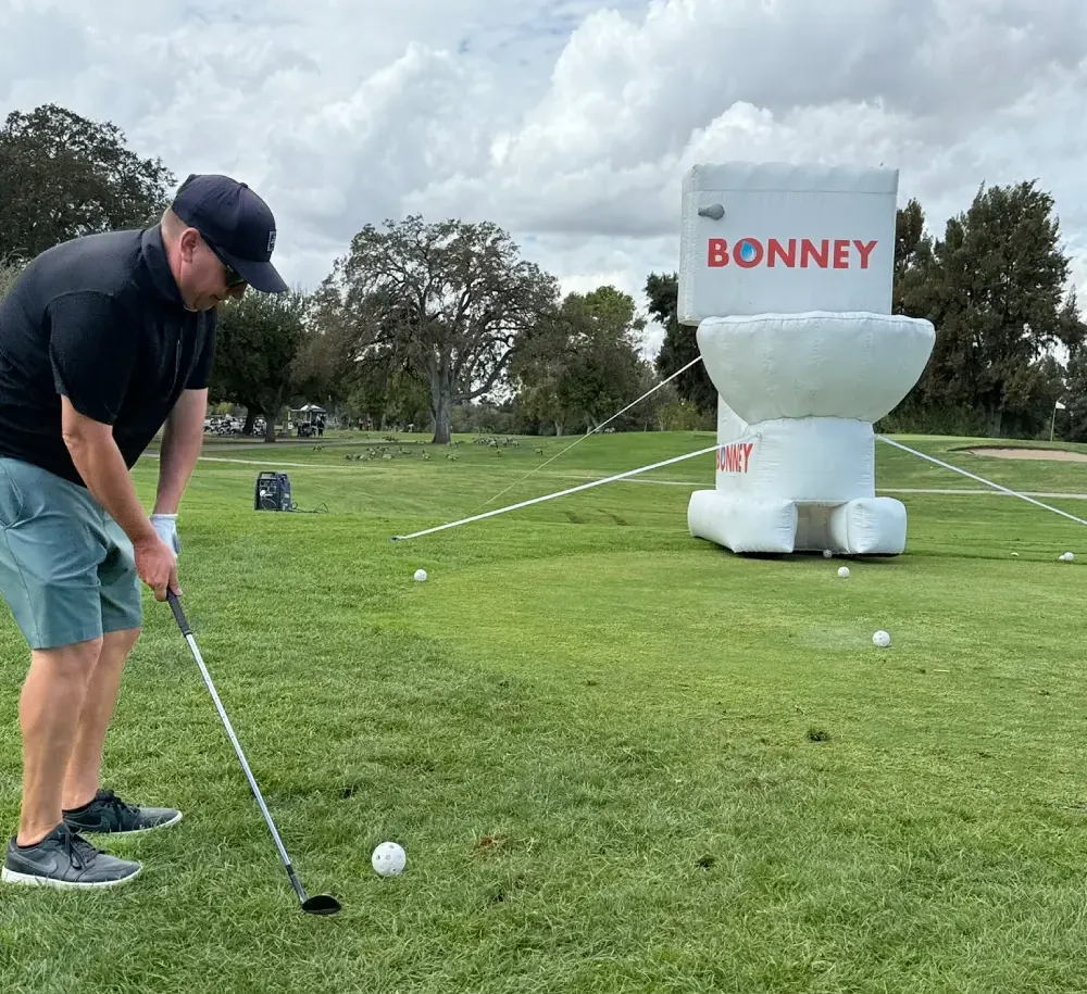 Man wearing a cap and black shirt swings a golf club on a grassy course toward a white Bonney inflatable target in the background.